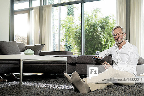 Älterer Mann mit grauen Haaren im modern gestalteten Wohnzimmer sitzt auf dem Boden und hält ein Tablett