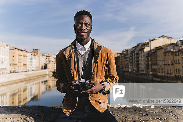 Porträt eines lächelnden jungen Mannes mit einer Kamera auf einer Brücke über den Arno  Florenz  Italien