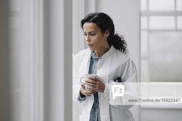 Ärztin steht am Fenster  sieht besorgt aus und hält eine Tasse Kaffee