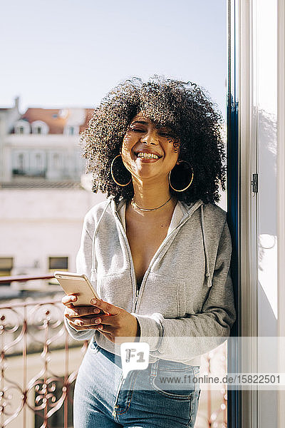 Porträt einer glücklichen jungen Frau mit lockigem Haar  die auf dem Balkon ihr Handy hält