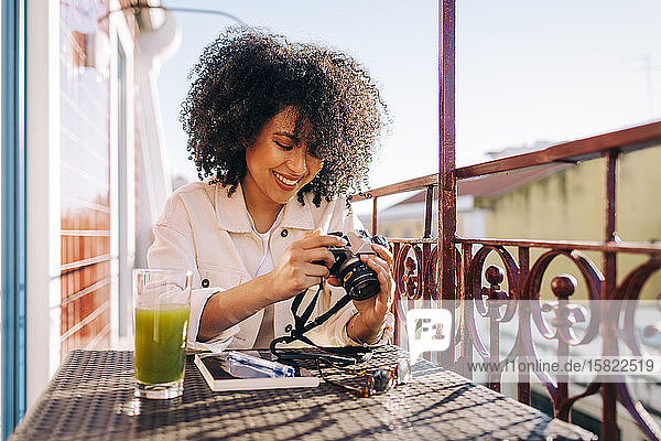 Lächelnde junge Frau mit lockigem Haar sitzt mit Smoothie und Kamera am Tisch auf dem Balkon