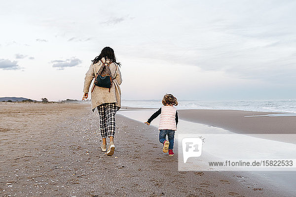 Rückansicht von Mutter und Tochter beim Laufen am Strand bei Sonnenuntergang