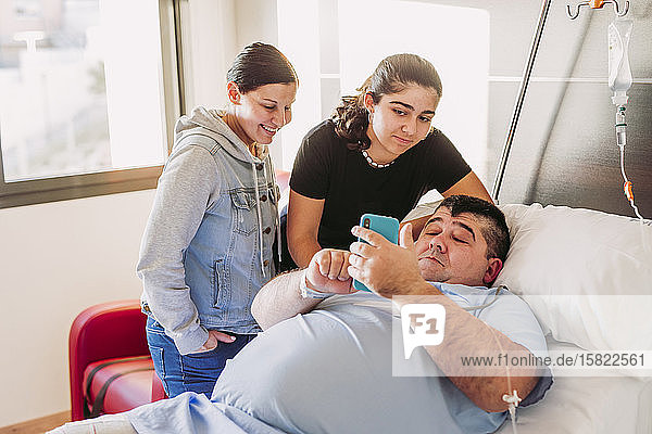 Familie besucht Mann mit Handy im Krankenhausbett liegend