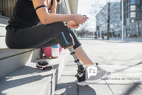 Sportliche junge Frau mit Beinprothese sitzt auf einer Treppe in der Stadt und benutzt ein Smartphone