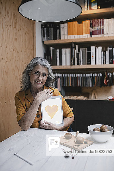 Lächelnde grauhaarige Frau hält Tablette mit einem Herz auf dem Bildschirm