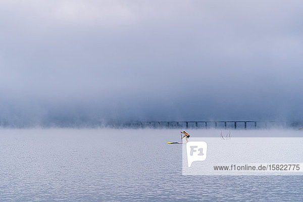 Mann steht beim Paddel-Surfen auf einem See im Nebel auf