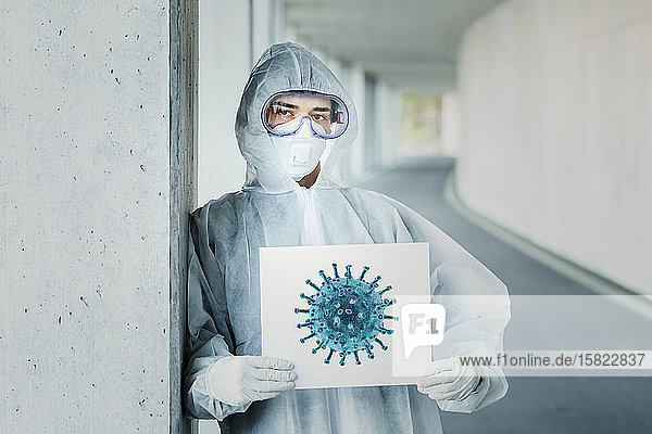 Porträt eines Mannes in Schutzkleidung der ein Schild mit der Darstellung eines Virus hält