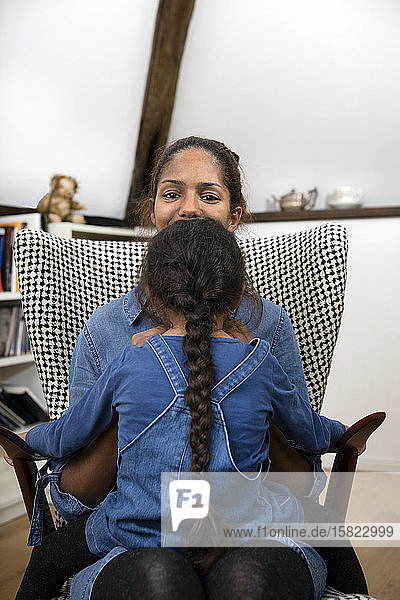 Back view of little girl with long braid sitting on her mother's lap on an armchair
