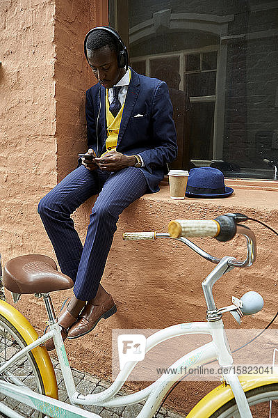 Stilvoller junger Geschäftsmann mit Fahrrad und altmodischem Anzug  der auf einem Fensterbrett sitzt und über Kopfhörer Musik hört