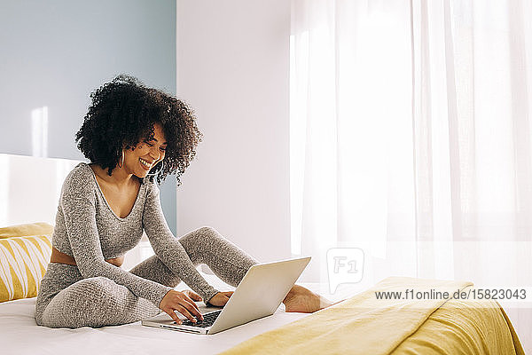 Happy young woman with curly hair using laptop on bed at home