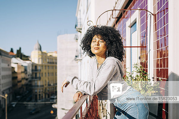 Porträt einer jungen Frau mit lockigem Haar  die auf einem Balkon steht und den Sonnenschein genießt