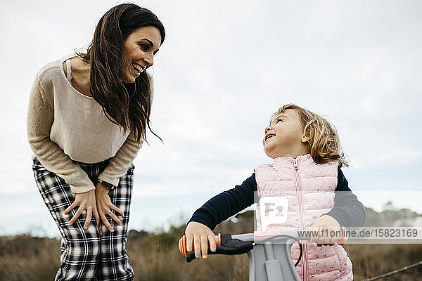 Happy mother looking at daughter with balance bicycle