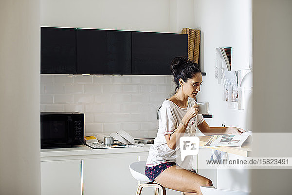 Junge Frau liest eine Zeitschrift und trinkt Kaffee in der Küche