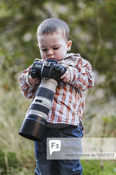 Porträt eines Kleinkindes  das eine digitale Spiegelreflexkamera mit großem Objektiv in der Natur betrachtet
