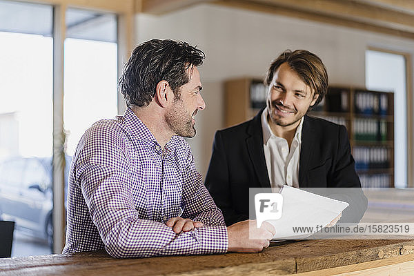 Zwei lächelnde Geschäftsleute mit Dokumenten im hölzernen Großraumbüro im Gespräch