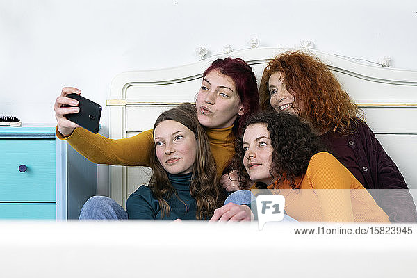 Mother and her three daughters sitting together on bed taking selfie with smartphone