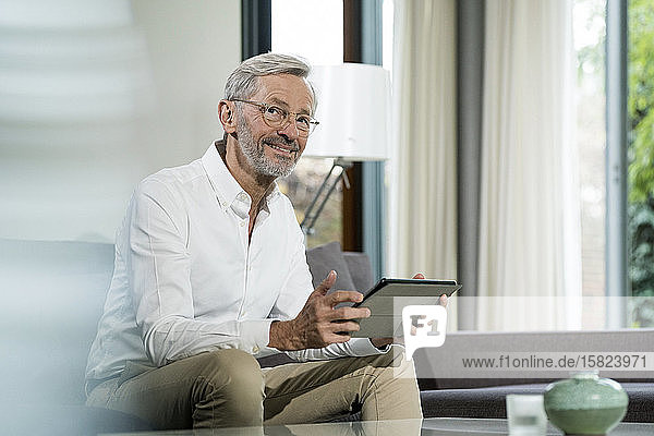 Lächelnder älterer Mann mit grauen Haaren im modern gestalteten Wohnzimmer  der auf einer Couch sitzt und ein Tablett hält