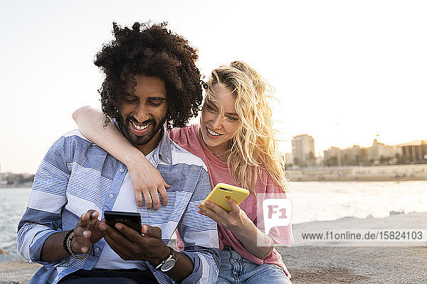 Glückliches junges Paar sitzt bei Sonnenuntergang mit einem Smartphone an der Kaimauer