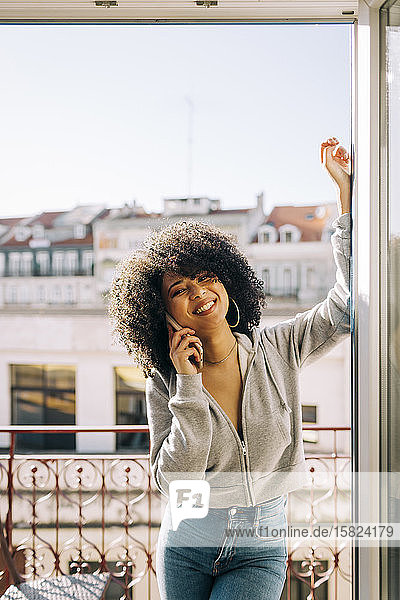 Porträt einer glücklichen jungen Frau mit lockigem Haar auf dem Balkon am Telefon