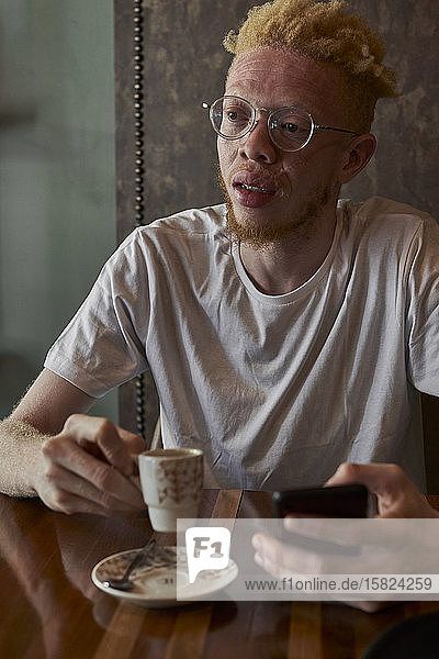 Albino man with round glasses having a coffee and checking his phone in a cafeteria