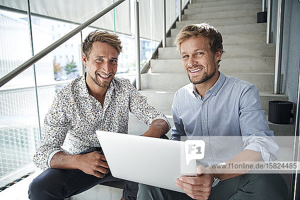 Porträt von zwei lächelnden jungen Geschäftsleuten  die mit Laptop auf einer Treppe sitzen