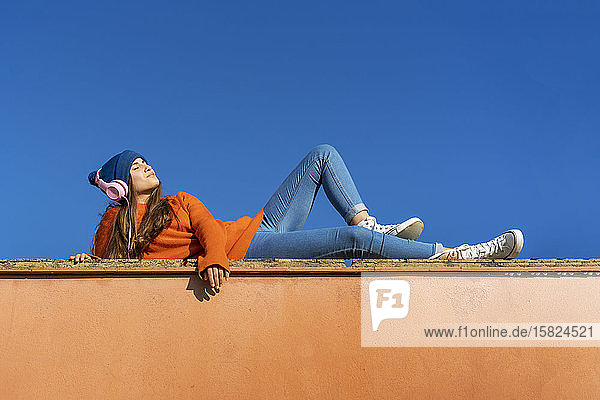 Teenager-Mädchen liegt an der Wand vor blauem Himmel und hört Musik mit Kopfhörern