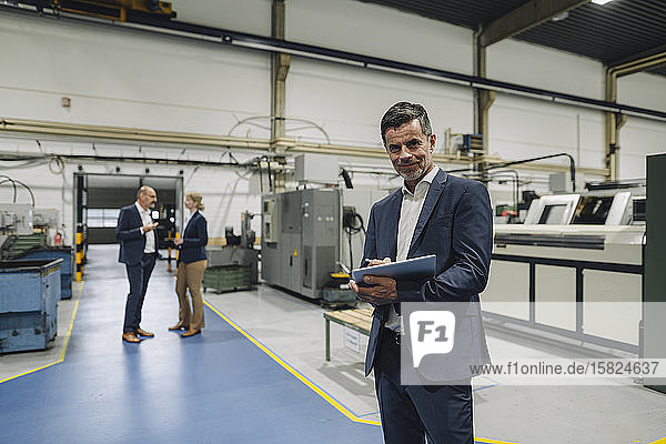 Porträt eines selbstbewussten Geschäftsmannes mit Tablette in einer Fabrik mit Kollegen im Hintergrund