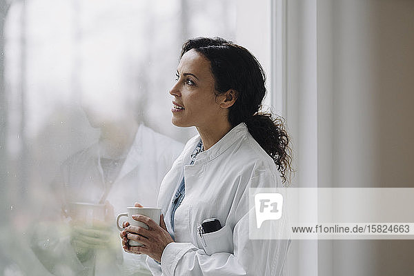Female doctor looking out of window  holding cup of coffee