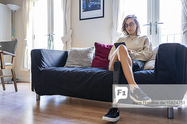 Junge Frau mit Beinprothese sitzt zu Hause auf Couch und liest E-Book