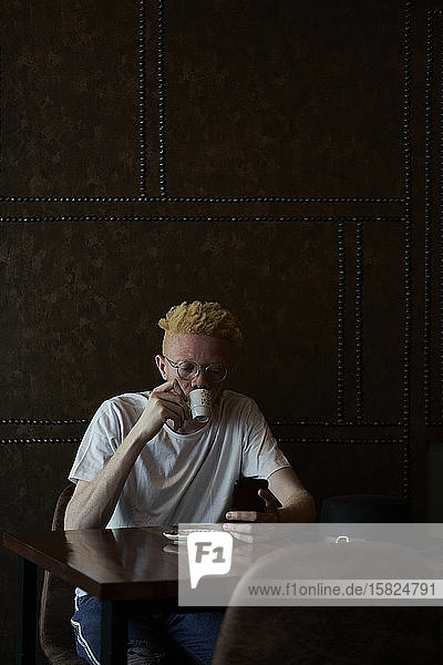 Albinomann mit runder Brille trinkt einen Kaffee und überprüft sein Telefon in einer Cafeteria