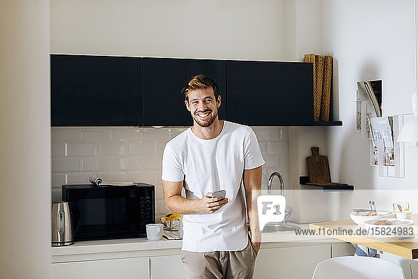 Porträt eines glücklichen jungen Mannes mit Handy in der Küche