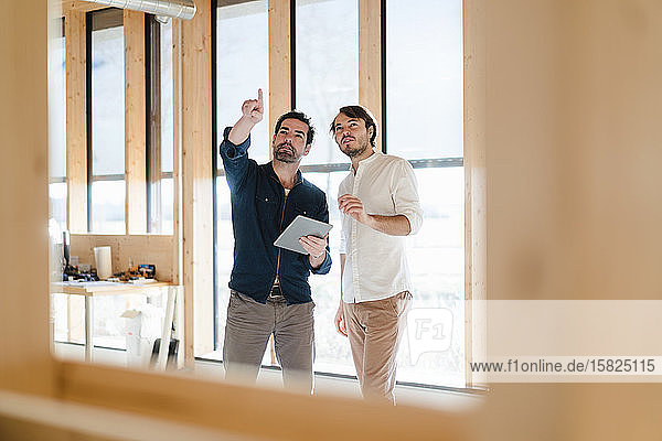 Zwei Geschäftsleute mit Tablett sprechen am Fenster im hölzernen Großraumbüro