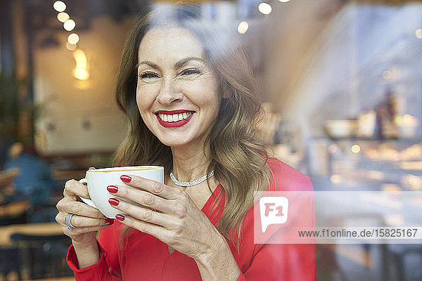 Porträt einer lachenden  reifen Frau mit einer Tasse Kaffee hinter einer Fensterscheibe