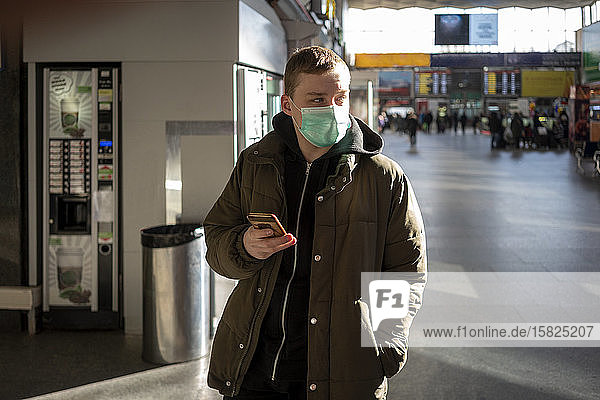 Junger Mann mit Gesichtsmaske am Bahnhof in der Stadt  der ein Smartphone in der Hand hält