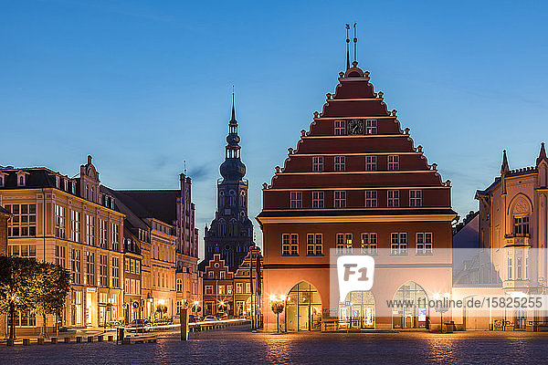 Deutschland  Mecklenburg-Vorpommern  Greifswald  Beleuchtetes Rathaus in der Abenddämmerung mit dem Turm des Heiligen Nikolai im Hintergrund