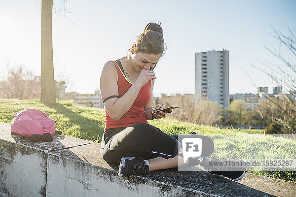 Sportliche junge Frau mit Beinprothese  die mit ihrem Handy an einer Wand sitzt