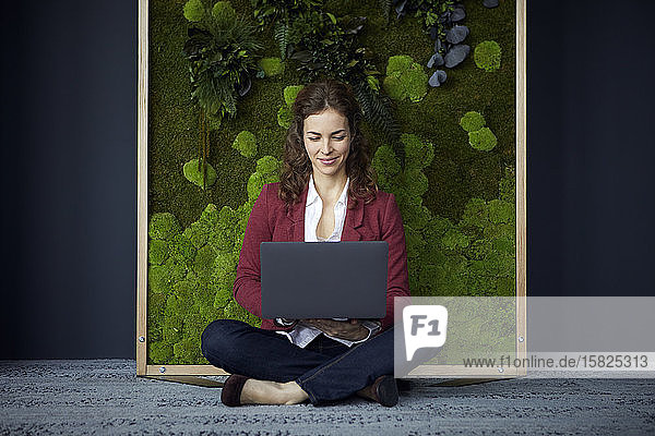 Smiling businesswoman sitting on the floor in green office using laptop