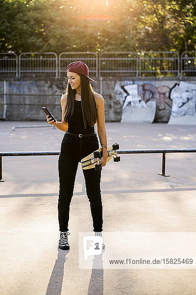 Junge Frau mit Smartphone und Skateboard im Skatepark
