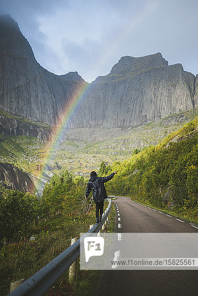 Norwegen  Lofoten-Inseln  Mann balanciert auf Leitplanke mit Bergen und Regenbogen im Hintergrund