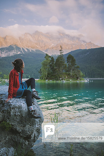 Deutschland  Bayern  Eibsee  Junge Frau sitzt auf einem Felsen und schaut auf die Aussicht am Eibsee in den bayerischen Alpen