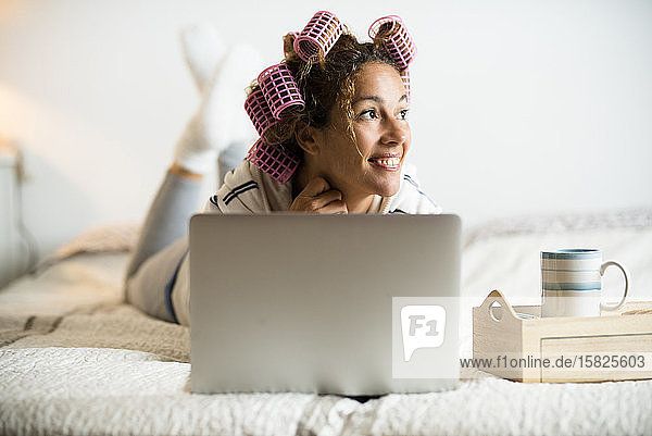 Woman with hair curlers lying on bed with laptop
