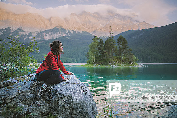 Germany  Bavaria  Eibsee  Young woman sitting on rock and looking at scenic view by EibseeÂ lake in Bavarian Alps