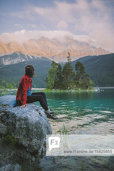 Germany  Bavaria  Eibsee  Young woman sitting on rock and looking at scenic view byÂ EibseeÂ lake in Bavarian Alps