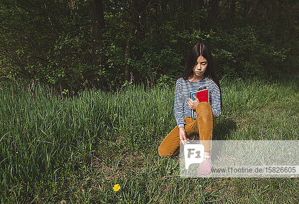 Ein nachdenkliches Mädchen sitzt im Frühling still mit einem Buch im hohen Gras