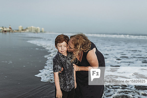 Sohn macht ein lustiges Gesicht  während Mutter ihn in der Abenddämmerung am Strand küsst