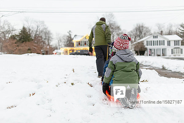 Rückansicht eines Vaters  der Kinder auf einem orangefarbenen Schlitten durch den Schnee zieht