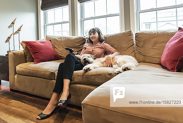 Young woman in business attire sitting on couch with dog watching tv.