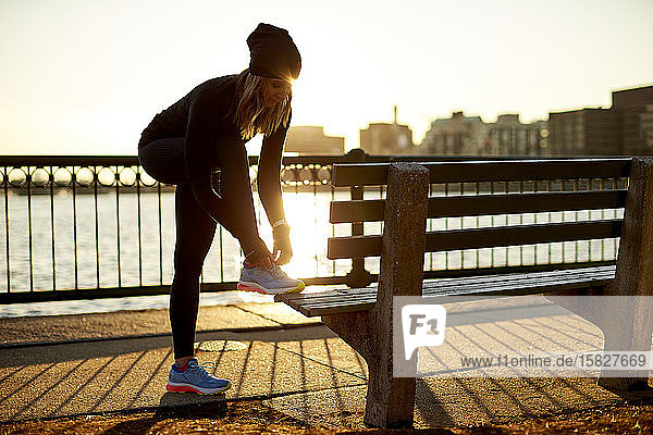 Eine von hinten beleuchtete Sportlerin bindet ihren Turnschuh auf einer Parkbank.