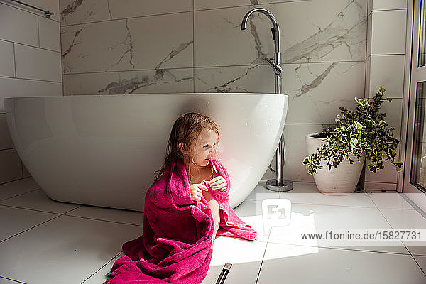 kleines Mädchen  das in einem rosa Handtuch auf dem Boden in der Badewanne sitzt
