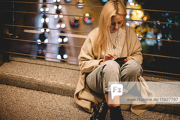 Junge Frau benutzt ein Smartphone  während sie nachts auf einer Brücke in der Stadt sitzt